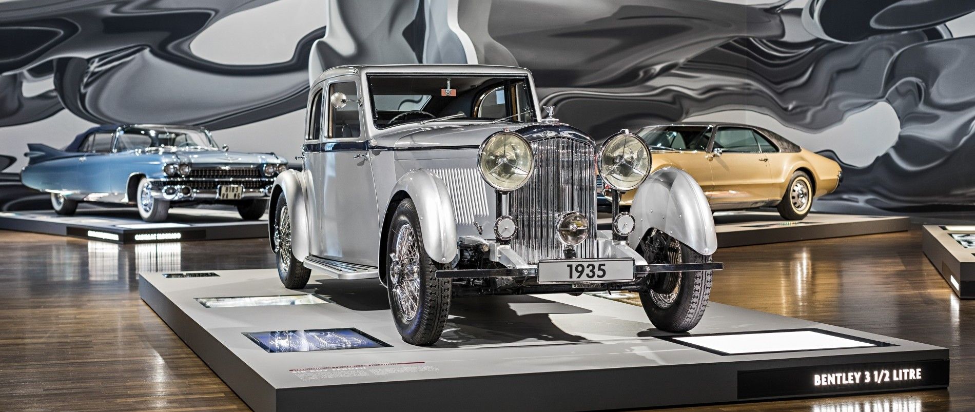 Klassische Autos in einem Ausstellungsraum, darunter ein Bentley 3 1/2 Litre von 1935, ein blaues Fahrzeug und ein goldenes Auto auf Podesten.