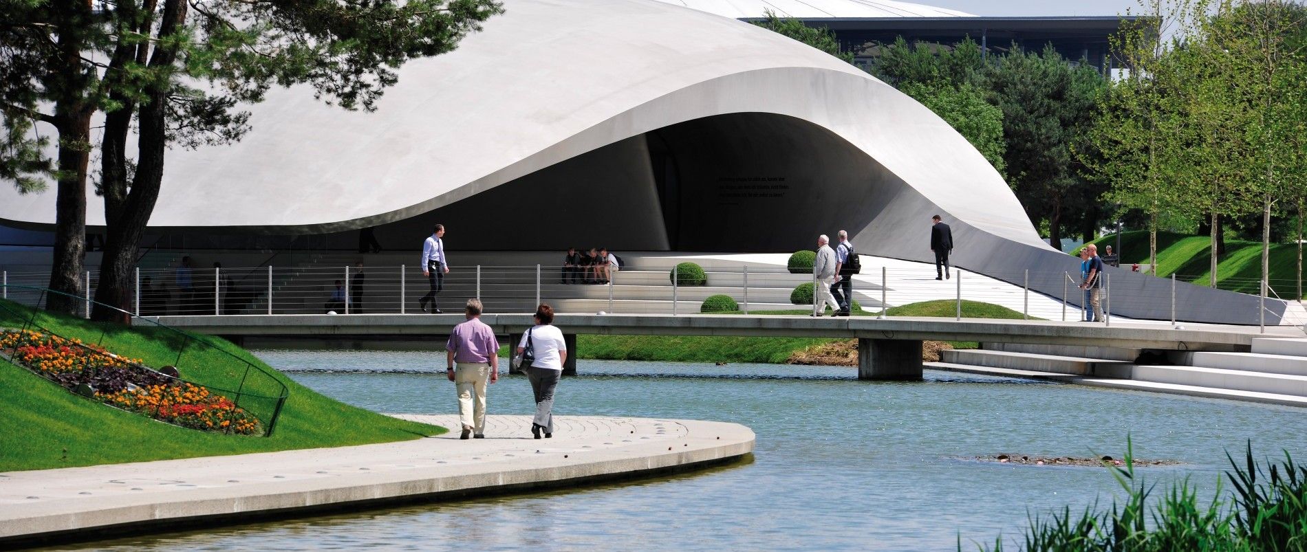 Moderne Architektur mit geschwungenem Gebäude und Menschen, die über Brücke gehen, umgeben von Wasser und Grünflächen.