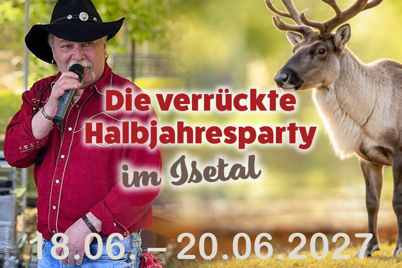 Ein Sänger im roten Westernhemd und schwarzen Cowboyhut steht mit Mikrofon neben einem Rentier in sommerlicher Naturkulisse.
