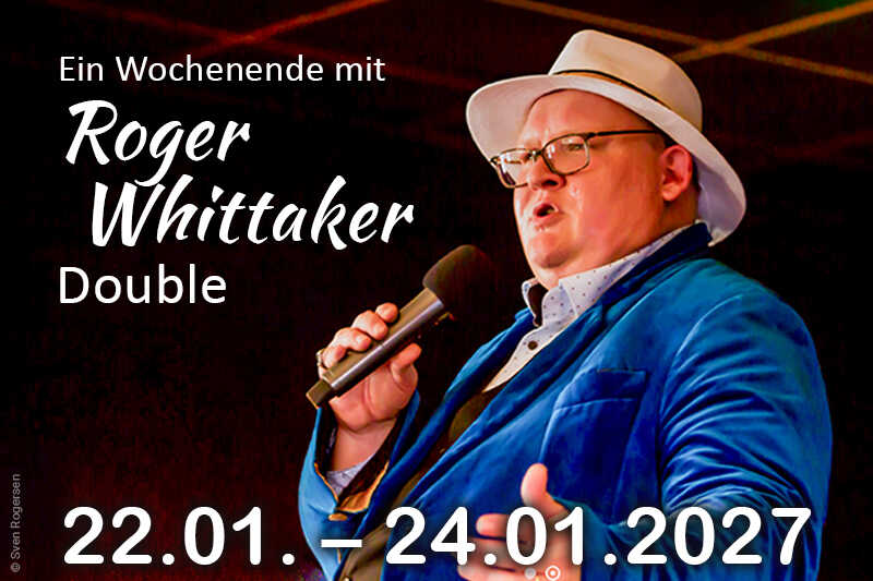 Ein Sänger im blauen Sakko und weißen Hut steht mit Mikrofon auf der Bühne vor einem dunklen, stimmungsvoll beleuchteten Hintergrund.