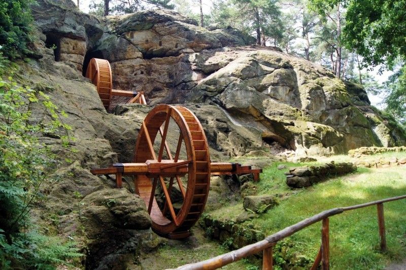Zwei große hölzerne Wasserräder an einem felsigen Hang in einem Waldgebiet.