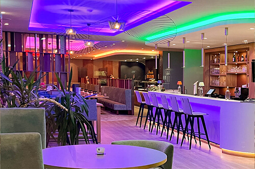 Moderne Bar-Lounge mit bunten LED-Leuchten, weißen Thekenstühlen und Pflanzen.