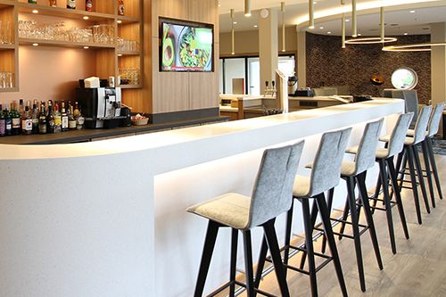 Moderne Bar mit hoher Theke, gepolsterten Barhockern und hinterleuchteten Regalen gefüllt mit Flaschen und Gläsern.