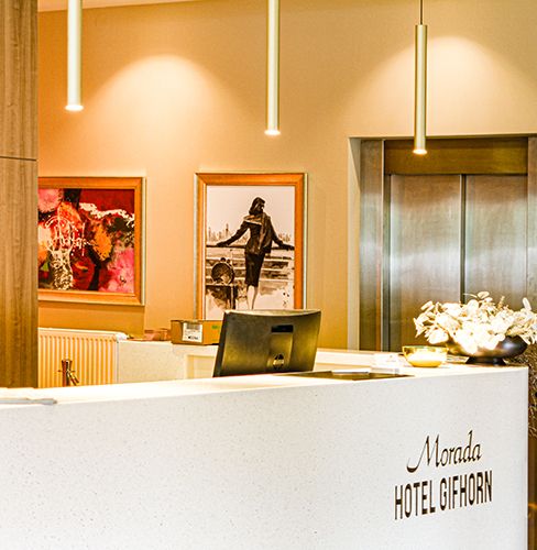 Empfangsbereich des Morada Hotels Gifhorn mit modernem Interieur, einem Tresen mit Computerbildschirm, dekorativer Blumenvase und zwei Kunstgemälden an der Wand.
