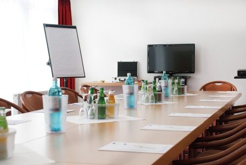 Konferenzraum mit langem Tisch, darauf Getränke, Gläser und Schreibmaterial. Im Hintergrund ein Bildschirm und ein Flipchart.