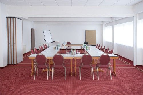 Ein leerer Besprechungsraum mit einem rechteckigen Tisch, umgeben von roten Stühlen. Der Raum hat einen roten Teppichboden und weiße Wände. Ein Flipchart und Erfrischungen stehen bereit.