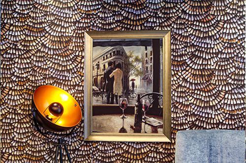 Wand mit dekorativem Muster, Gemälde eines Paares in einer Straßenszene, daneben eine orange beleuchtete Stehlampe.