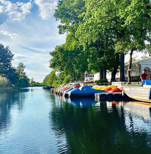 Wasserkanal mit Bäumen, parallel angelegte bunte Tretboote und ein größeres Schiff an einem sonnigen Tag.
