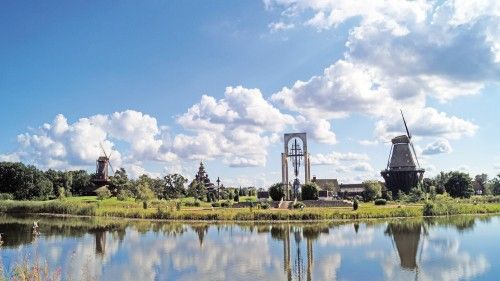 Landschaft mit zwei Windmühlen und einer Kirche am Ufer eines Sees mit blauem Himmel und Wolken.