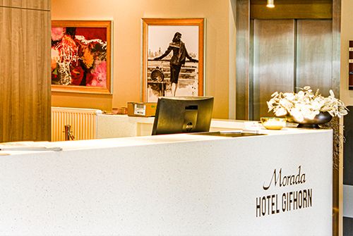Empfangsbereich des Morada Hotel Gifhorn mit Theke, Kunstwerken an der Wand und Computer am Tresen.