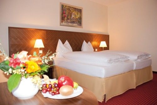 Doppelbett in einem Hotelzimmer mit Nachttischlampen, Blumenstrauß und Obstteller im Vordergrund.