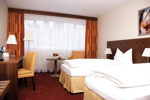 Modern eingerichtetes Hotelzimmer mit zwei Einzelbetten, einem Schreibtisch, Sesseln und einem kleinen Tisch, helle Vorhänge und warme Farbgestaltung.