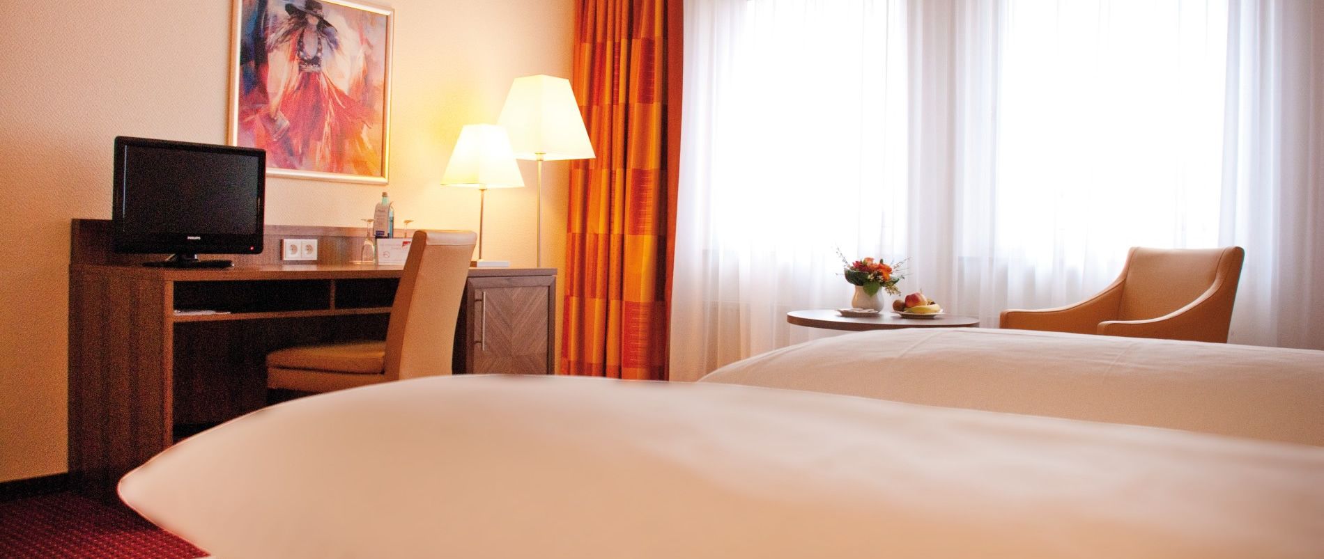 Hotelzimmer mit zwei Betten, Schreibtisch mit Fernseher, Sessel und Tisch mit Blumenvase vor einem Fenster.